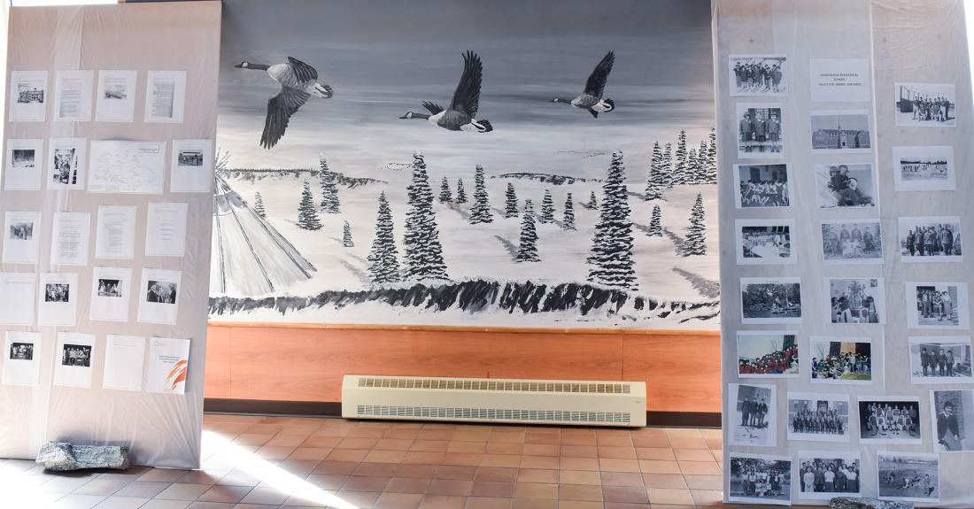 Fort George residential school photos in front of mural of 3 flying geese